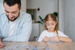 © Julija - Smart game. Man and his daughter sitting at a table and looking at puzzle pieces while playing at home together. Focus on girl