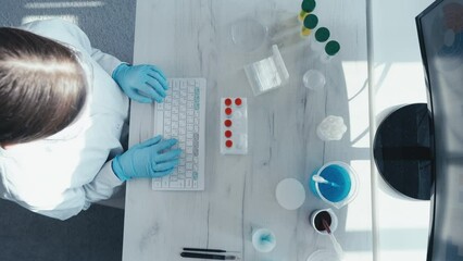 Wall Mural - A female researcher sits at a workplace in a laboratory, behind a personal computer monitor. Against the background research statistics. Pharmaceutical medical worker in protection works at keyboard.