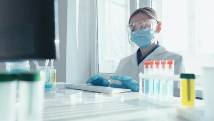 Aufkleber - A female researcher sits at a workplace in a laboratory, behind a personal computer monitor. Against the background research statistics. Pharmaceutical medical worker in protection works at keyboard.