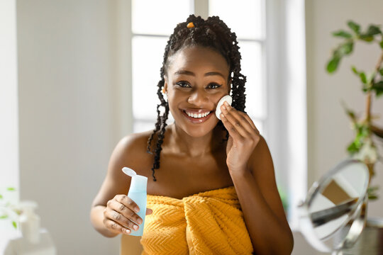 Wall Mural - Face care routine. Happy young black woman looking at camera, using micellar water and cotton pad for makeup removal