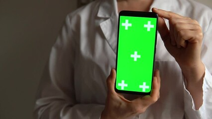 Woman doctor holding green screen chroma key smartphone in clinic. Layout for mobile applications. Female in medical uniform show app on phone screen. Concept of programs and advertising for medicine