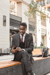 Sticker - Portrait of black african american businessman in suit with glasses and headphones sits on the background of city buildings outdoors