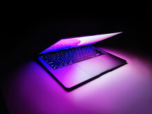 Laptop With Half Lid Open On A Table Lit With Colorful Purple And Blue Desktop Screen Wallpaper In A Dark Room