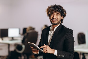 Wall Mural - Excited businessman scrolls tablet screen. Indoor shot of smiling entrepreneur with digital device. Happy office worker looking at camera