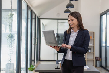 Poster - A stylish white businesswoman in a black suit standing inside the office in hands with a mobile phone, tablet, and laptop waiting for customers to contact, business people concept.