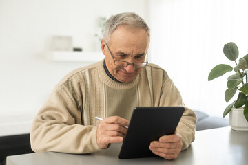 Senior man reading news on digital tablet. Serious mature male using portable computer at home, sitting at table, copy space.