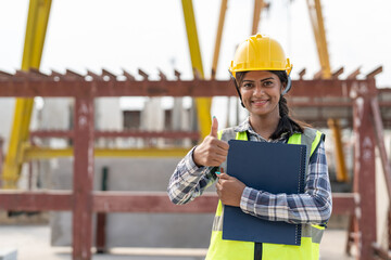 Asian woman engineer holding document smiling at construction site. Confident female Indian wearing protective helmet and vest working in factory making precast concrete wall for real estate housing.