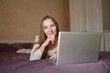 © GRON777 - Young woman in a country house, working on a laptop, lying on the bed, watching online entertainment, smiling, free time at home. Technology concept, country rest