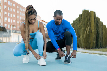 A dark-haired couple spends a happy afternoon playing sports outdoors, the black man and the black woman with braids bend down to tie their shoes. Concept of sport as a couple.