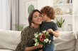 © New Africa - Little son kissing and congratulating his mom with Mother`s day at home. Woman holding bouquet of flowers