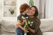 Little Son Kissing And Congratulating His Mom With Mother`s Day At Home. Woman Holding Bouquet Of Flowers