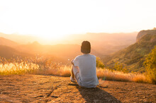Man Sitting On Top Of A Mountain While Watching The Sunset In The Background. Person Having Reflections And Thoughts While Contemplating The Landscape.