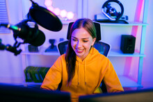 Young Caucasian Woman Professional Gamer Wearing Yellow Hoodie Sits On A Chair With A Gaming Table With Pc, Keyboard, Monitor, Microphone. Prepare For Competition, Cast Gameplay Or Record A Podcast.