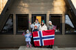 Portrait of family with kids outside cabin house holding Norway flags. Scandinavian culture, norwegian people.