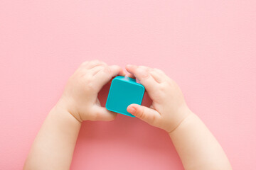 Baby girl hands holding and playing with light blue plastic shape on light pink table background. Pastel color. Closeup. Infant development toy. Point of view shot. Top down view.