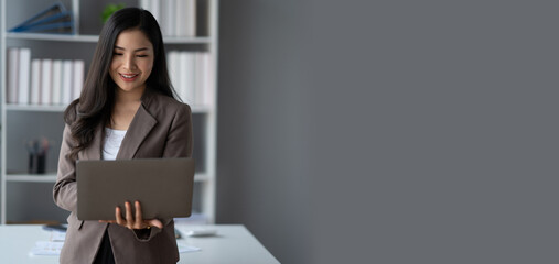 Beautiful Asian businesswoman using laptop computer while standing in the modern office room.