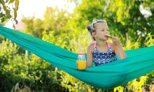 Portrait Of Happy Kid Cute Girl Is Eating Pizza And Drink Orange Juice From Jar With Straw Sitting On Green Hammock On Warm Day. Summer Holiday Vacation.
