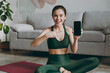 © ViDi Studio - Smiling young strong sporty athletic fitness trainer instructor woman wear green tracksuit use blank screen mobile cell phone training do exercises at home gym indoor Workout sport motivation concept