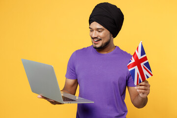 Poster - Smiling fun devotee Sikh Indian IT man ties his traditional turban dastar wear purple t-shirt hold use work on laptop pc computer hold British flag isolated on plain yellow background studio portrait.