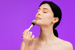 © wedmoments.stock - Close up portrait of attractive beautiful young topless woman standing isolated over purple background holding makeup brush