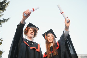 Portrait of happy graduates. Two friends in graduation caps and gowns standing outside university building with other students in background, holding diploma scrolls, and smiling