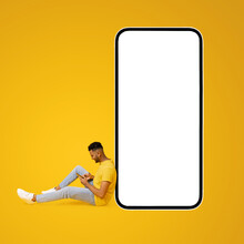 Handsome Indian Man Sitting Beside A Large, Oversized Smartphone With Blank Screen, Copy Space And Mockup For Other Digital Content, Guy Is Using Mobile Phone To Send Text Messages Or Browse Online