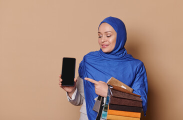 Pleasant Arab Muslim female sales manager in blue hijab, holds fabric samples, shows smartphone with blank black screen with space for mobile application or advertising, isolated beige background