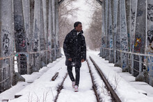 African American Man Walking On Railway Track In Bridge Snowy Winter In Countryside Forest Outside, Falling Snow. Positive Black Man Going Outdoors. Leisure Activity Concept. Copy Text Space For Ad