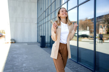 Excited businesswoman reading good news on smartphone while standing outdoors. Happy lady worker in suit making yes gesture with office center on background.