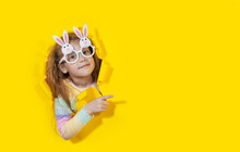 Cute  Child Girl In Bunny Shape Glasses Looking, Peeping Through The Yellow Paper Hole. Advertise Childrens Goods For Easter Holidays. Copy Space For Text. Pointing To The Right