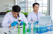 © Bangkok Click Studio - Asian professional male scientist researcher in white lab coat and rubber gloves sitting using microscope inspecting quality of vegetable in laboratory while colleague typing data in laptop computer