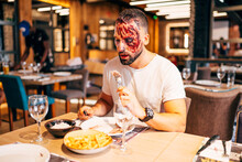 Man With Makeup On His Face With Wounds And Blood. Eating At The Table