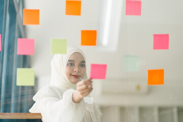 A Muslim businesswoman uses Post-it notes to share ideas, showcasing the concept of brainstorming. The sticky notes are placed on a glass wall.