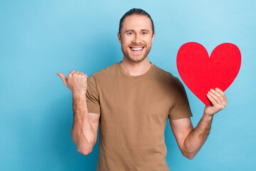 Sticker - Photo of cheerful positive handsome guy dressed beige t-shirt indicating empty space hand hold heart isolated on blue color background