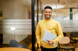 Smiling young indian businessman, arab male student, eastern employee, manager, coworker wearing casual yellow pullover standing carrying laptop, looking at the camera standing in office