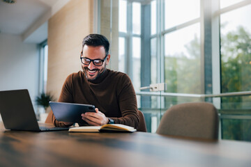 Smiling businessman using a digital tablet, sitting in front of the laptop at the office.