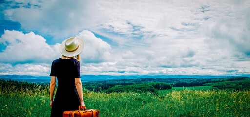 Sticker - Blonde woman in black dress and suitcase at meadow with mountains on background