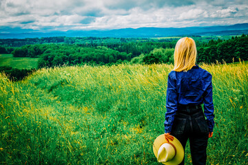 Wall Mural - Blonde cowgirl in hat at meadow with mountains on background