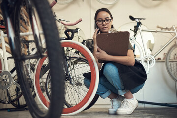 Wall Mural - Is it the white or pink one. Full length shot of an attractive young woman crouching in her bicycle shop and using her cellphone while holding a clipboard.