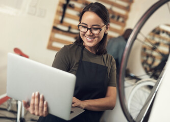 Wall Mural - Emails are how I stay in touch with clients. Shot of an attractive young woman standing alone in her bicycle shop and using her laptop.