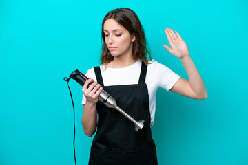 Young caucasian cooker woman using hand blender isolated on blue background making stop gesture and disappointed