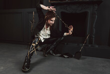 Portrait Of A Mystical Or Magical Girl With A Gothic Make-up And Auburn Hair, Dressed In A Long Coat, Off-white Shirt, Tight Leather Leggings And Tall Laced Chunky Boots. Posing In A Dark Gothic Room.