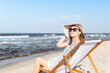 © rogerphoto - Happy brunette woman wearing sunglasses and hat relaxing on a wooden deck chair at the ocean beach.