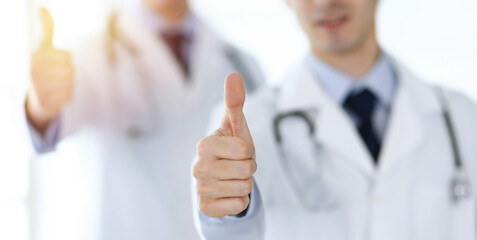 Two male doctors standing as a team and showing thumbs up as a symbol of the best service for patients in sunny clinic. Medicine concept