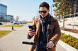© baranq - Sustainable commuting. Handsome man using smartphone while going to work on electric scooter