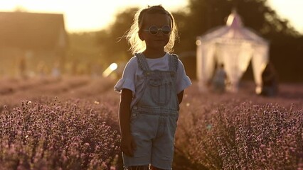 Wall Mural - Adorable little girl is having fun against the background of a large lavender field on sunset. Hyperactive smiling little kid in sunglasses on nature. International Children's Day.