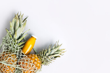 Two ripe juicy pineapples and natural fresh pineapple juice in a glass bottle on a gray background in a green string bag. Healthy, detox and dietary nutrition concept.