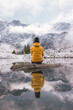 person in yellow jacket reflection in a calm pond in the mountains with snowy mountains in distance