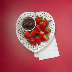 Wall Mural - Top View of Ripe Strawberries on a Heart-Shaped Plate with Chocolate Dipping Sauce in a Bowl on a Red Background