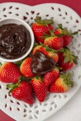 Wall Mural - Close-up of Fresh Strawberries on a Heart-Shaped Plate on Red Background; Bowl of Melted Chocolate for Dipping; Chocolate-Dipped Strawberry on Top